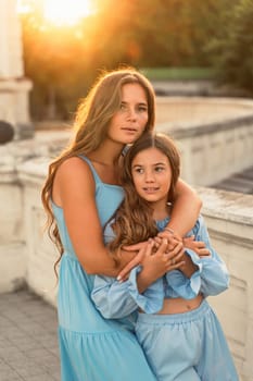 Portrait of mother and daughter in blue dresses with flowing long hair against the backdrop of sunset. The woman hugs and presses the girl to her. They are looking at the camera