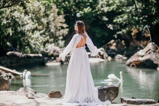 a beautiful woman in a long white dress looks into the distance at a beautiful lake with swans