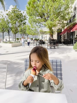 Little girl licks popsicles in a waffle cone while sitting on a chair in a street cafe. High quality photo
