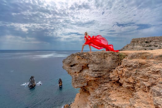 A woman in a red silk dress stands by the ocean, with mountains in the background, as her dress sways in the breeze