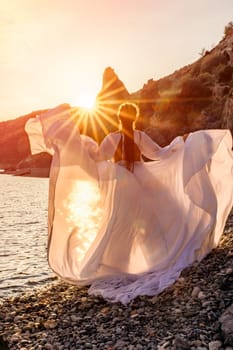 Mysterious woman silhouette long hair walks on the beach ocean water, sea nymph wind listens to the wave. Throws up a long white dress, a divine sunset. Artistic photo from the back without a face.