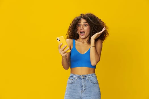 Influencer woman with phone in hand taking selfies talking on video call freelancer social media technology communication at work, with curly afro hair in blue t-shirt on yellow background , smile with teeth and happiness, copy space. High quality photo