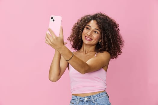 Woman blogger holding phone in hand video call, with curly hair in pink top and jeans poses on pink background, copy space, technology and social media, online. High quality photo