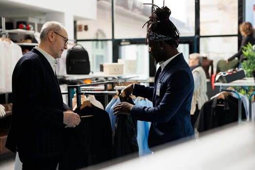 African american employee helping customer with suit in shopping centre, client looking to buy modern clothes. Elderly man buying fashionable merchandise in showroom. Fashion concept