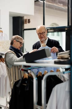 Senior couple looking at blue trendy shirt analyzing material in clothing store, looking at new fashion collection. Elderly people shopping for casual wear, buying fashionable merchandise