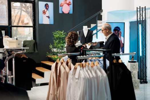 Showroom employee helping senior man choosing right tie for formal wear in modern boutique, discussing accessories fabric. Elderly client buying fashionable merchandise in clothing store