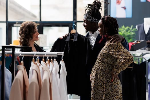 Worker helping diverse couple choosing trendy black shirt for casual wear in shopping centre. Clients discussing clothes fabric with employee before buying fashionable merchandise in modern boutique