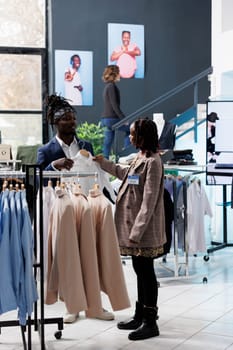 Boutique worker helping stylish customer with choosing white shirt during commerical activity. African american client buying fashionable clothes and accessories in shopping centre. Fashion concept