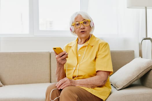 Happy elderly woman looking into her phone video call with headphones smile, technology for communication, bright modern interior, lifestyle online communication. High quality photo