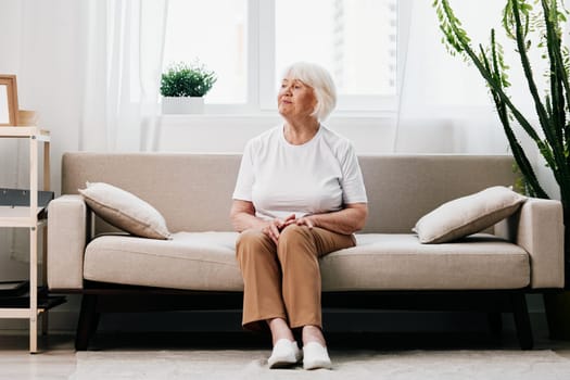 Elderly woman sits on sofa at home, bright spacious interior in old age smile, lifestyle. Grandmother with gray hair in a white T-shirt and beige trousers. High quality photo