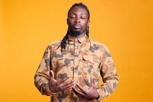 African american man taking deep breaths, meditating in studio over yellow background. Concentrated young adult relaxing during body muscles peaceful activity, enjoying practicing healthy lifestyle