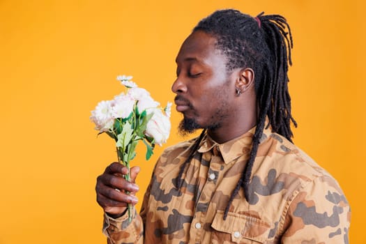 African american man smelling white roses bouquet, posing with flowes for valentine s day in studio over yellow background. Smiling romantic young adult enjoying love holiday