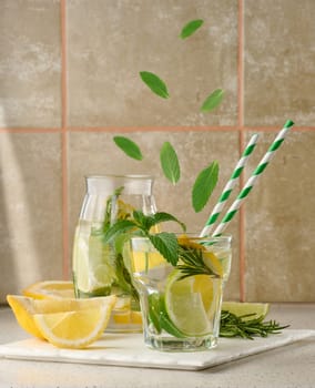 Lemonade in a transparent glass with lemon, lime, rosemary sprigs and mint leaves on a white background