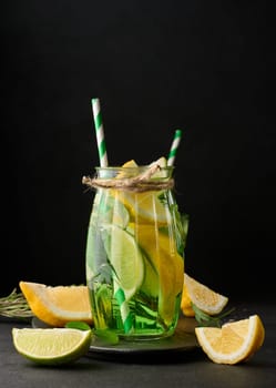 Lemonade in a transparent glass with lemon, lime, rosemary sprigs and mint leaves on a black background