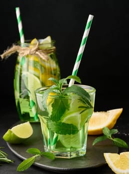 Lemonade in a transparent glass with lemon, lime, rosemary sprigs and mint leaves on a black background