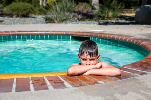 Concept of fun, health and vacation. Oudoor summer activity. A sad boy eight years old in swimming goggles is holding onto the side of the pool on a hot summer day.