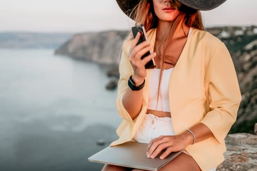 Successful business woman in yellow hat working on laptop by the sea. Pretty lady typing on computer at summer day outdoors. Freelance, travel and holidays concept.