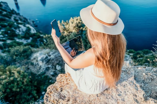 Successful business woman in yellow hat working on laptop by the sea. Pretty lady typing on computer at summer day outdoors. Freelance, travel and holidays concept.