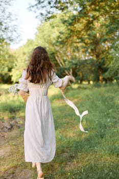 a woman in a long summer dress walks along a forest path holding a wicker hat with flowing ribbons in her hands. High quality photo