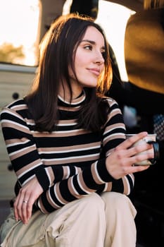 young caucasian woman smiling happy sitting in a camper van drinking a cup of coffee at sunset, concept of van life and weekend getaway