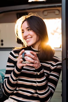 young woman smiling happy sitting in a camper van drinking a cup of coffee at sunset, concept of van life and weekend getaway