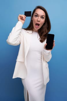 surprised brunette young woman in a dress and jacket with a payment card for shopping and a smartphone.