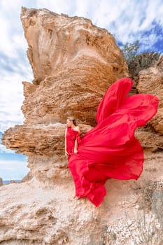 A woman in a red silk dress stands by the ocean, with mountains in the background, as her dress sways in the breeze