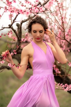 Woman peach blossom. Happy curly woman in pink dress walking in the garden of blossoming peach trees in spring.