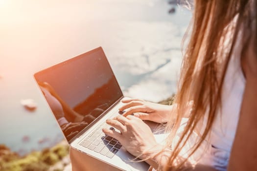 Successful business woman in yellow hat working on laptop by the sea. Pretty lady typing on computer at summer day outdoors. Freelance, travel and holidays concept.