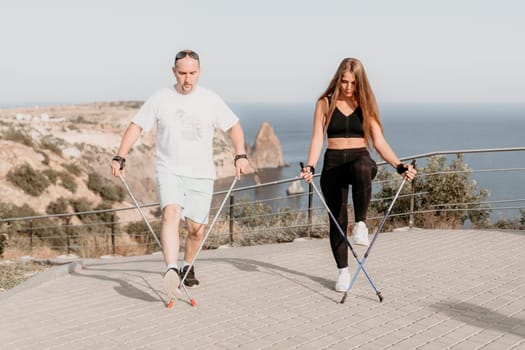 Happy Middle aged couple or friends practicing nordic walking in park near sea. Mature couple with trekking poles walking, practicing Nordic walking outdoors. Aging youthfully and sport concept.