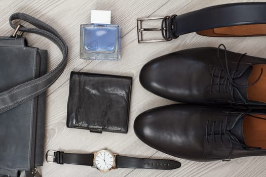 Pair of black leather men's shoes, leather shoulder bag for men, men's cologne, watch, belt and purse on gray wooden background. Men's accessories. Top view.