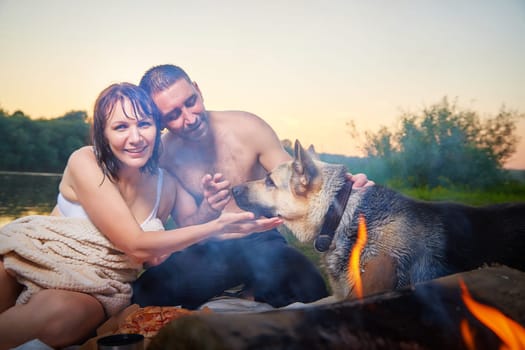 Happy wet couple relaxin, having fun and hugs with big dog near fire in camping on nature in summer sunny evening in sunset. Family or lovers have date and rest outdoor. Concept of love