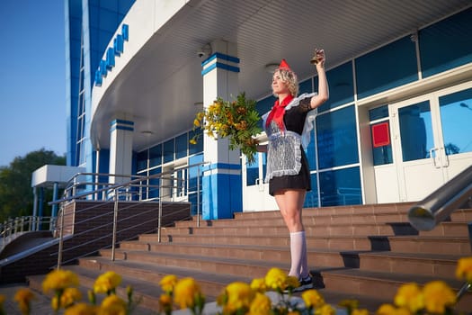 A Girl in black uniform, white apron, red tie on steps of school with bouquet of flowers and bell. Nostalgia photo shoot of teenager of female pioneer from USSR costume for September 1 or graduation
