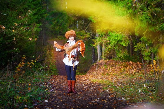 Girl in a leather jacket, a big red fox fur hat and with the skin of a fox killed on the hunt in the forest in autumn. A female model poses as fabulous royal huntress on nature hunt at photo shoot