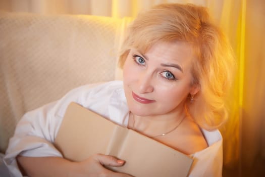 Adult mature woman of 40 or 60 years old with book in casual dress white shirt and blue jeans in calm cozy evening atmosphere room. Interior with transparent curtains and soft warm lamp