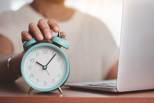 Asian woman's hand reaching for an alarm clock to check the time on desk with a computer laptop for the concept of work, study and time management.