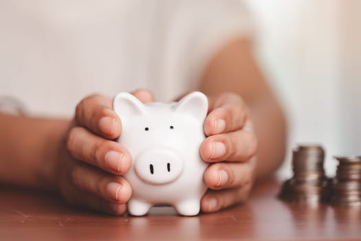 Asian women's hand holding a piggy bank with blurred coins and money on the wooden table for investment, business, finance and saving money concept.