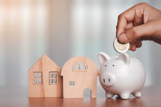 Asian women's hand saving a coin into piggy bank with wooden house model on the table for business, finance, saving money and property investment concept.