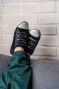 The child is lying in classic sneakers on a cozy sofa in the living room, with his feet on the back of the sofa, resting and relaxing. comfortable casual clothes and shoes. Relaxed legs of a boy in green casual pants and comfortable sneakers on a cozy sofa in the living room against a light brick wall