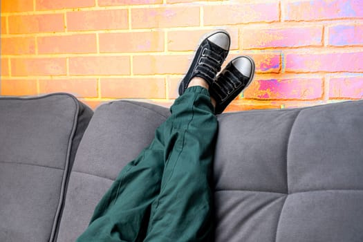 Relaxed legs of a boy in green casual pants and comfortable sneakers on a cozy sofa in the living room against a neon orange brick wall. children's feet in fashionable sneakers lie on the back of the sofa. Space for copying. Light Orange neon brick background
