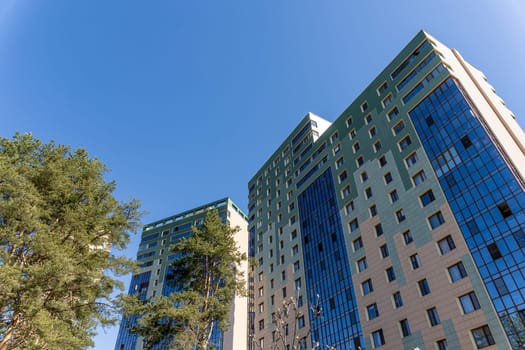 a tall high-rise building finished with glass. Beautiful apartment building on the background of blue sky and green pines. New residential neighborhood