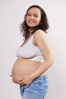 Vertical studio portrait: attractive happy pregnant multi ethnic woman holding hands on her tummy, smiling with beautiful toothy smile, looking at camera, isolated white background. Pregnancy fashion