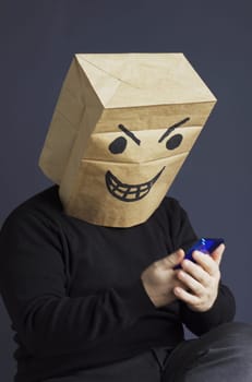 A man in a black turtleneck with a paper bag with an angry emoji on his head is typing a message on his smartphone while sitting on a chair. Emotions and anger. Vertical frame.