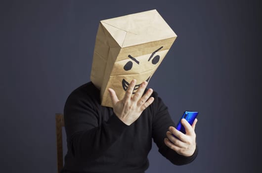 A man in a black turtleneck with a paper bag with an angry emoticon on his head is talking on a video link on a smartphone, gesturing with his hand. Emotions and anger
