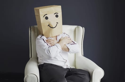 A man in a white shirt with a paper bag on his head, with a drawn smiley face, sits in an armchair and expresses his feelings - holding on to his heart.