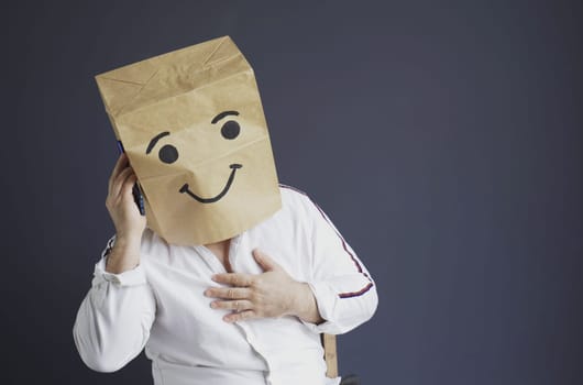 A man in a white shirt with a paper bag on his head, with a smiley face drawn, talking on the phone.