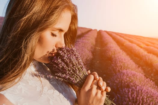 Close up portrait of young beautiful woman in a white dress and a hat is walking in the lavender field and smelling lavender bouquet.