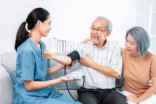 An elderly man having a blood pressure check by his personal caregiver with his wife sitting next to him in their home.