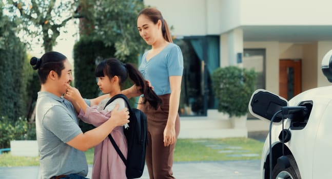 Progressive young parents and daughter with electric vehicle and home charging station. Green and clean energy from electric vehicles for healthy environment. Eco power from renewable source at home.