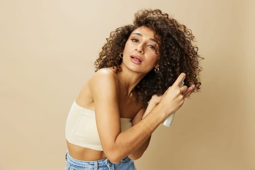 Woman applies cream and balm to her curly hair, the concept of protection and care with salon products, a healthy look, a smile with teeth on a beige background. High quality photo